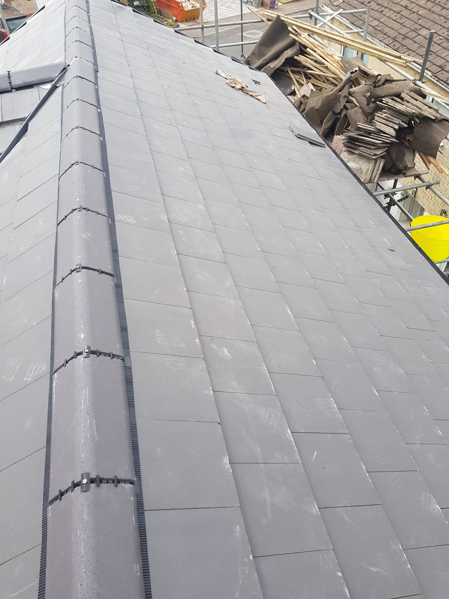 Newly finished anthracite smooth-tiled pitched roof with a clip-fixed dry ridge, stripped old tiles stacked on the scaffold behind